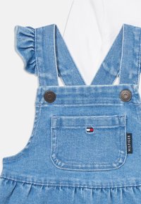Lys blå denim babyoverall med volanger på skulderstroppene, frontlomme med liten Tommy Hilfiger-logo og merkelapp på siden.