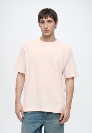 Jeune homme aux cheveux courts et bruns, portant un t-shirt oversize rose clair et un jean bleu, debout devant un fond blanc uni.