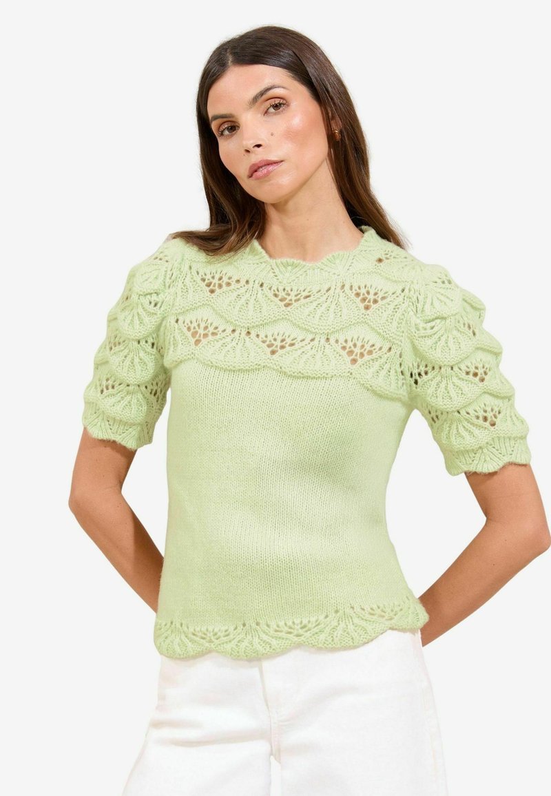 Femeie îmbrăcată cu un top tricotat de culoare verde deschis, cu modele de dantelă cu margini ondulate pe mâneci și piept, asortat cu pantaloni albi, pe un fundal simplu.