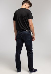 Młody mężczyzna w czarnej koszulce, ciemnoniebieskich dżinsach i granatowych trampkach, stojący plecami do widza na szarym, jednolitym tle.