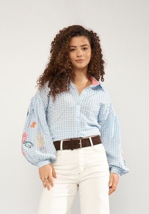 Noor naine lokkis juustega kannab sinist ruudulist pluusi, millel on tikitud mereloomad puffy varrukatel, ja valgeid pükse pruunis vöös.