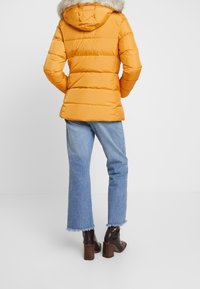 Person med en ljus orange pufferjacka med pälsmössa, ljusblå korta jeans med råa kanter och mörkbruna klackstövlar.