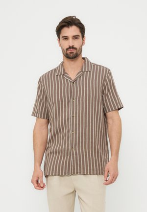 Άνδρας που φοράει κοντομάνικο πουκάμισο με κατακόρυφες ρίγες σε καφέ και λευκό χρώμα και ανοιχτό μπεζ παντελόνι, στέκεται μπροστά σε ένα απλό φόντο.