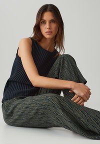 Top a canottiera nera a coste e pantaloni larghi verdi con un motivo a foglie. Modella seduta, gambe incrociate, con capelli arruffati e sfondo neutro.