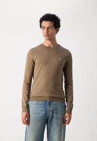 Bruine gebreide trui met een ronde hals, lange mouwen en geribde manchetten; heeft een klein blauw logo op de borst. Draagt men met lichtblauwe spijkerbroek.