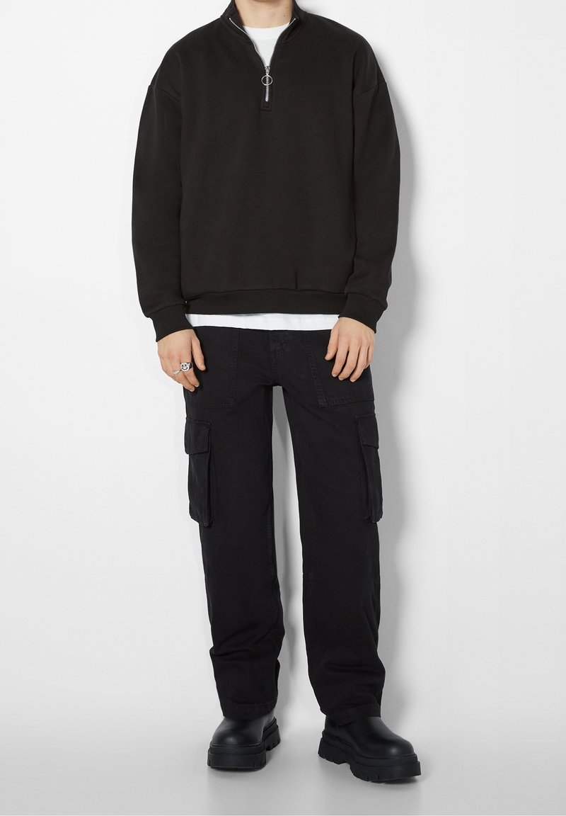 Svart zip-up sweatshirt i mjukt material, kombinerad med svarta cargobyxor med sidofickor och kraftiga svarta stövlar.