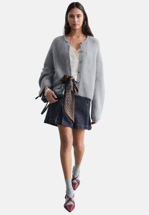 Kobieta ubrana w szary, puszysty kardigan, biały koronkowy top, ciemną dżinsową spódnicę, szare skarpetki, bordowe szpilki, trzymająca czarną torbę z ozdobną apaszką.