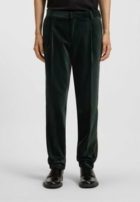 Grüne Samenhosen mit glatter Textur, ausgestattet mit einer taillierten Passform und Falten, kombiniert mit schwarzen formellen Schuhen.