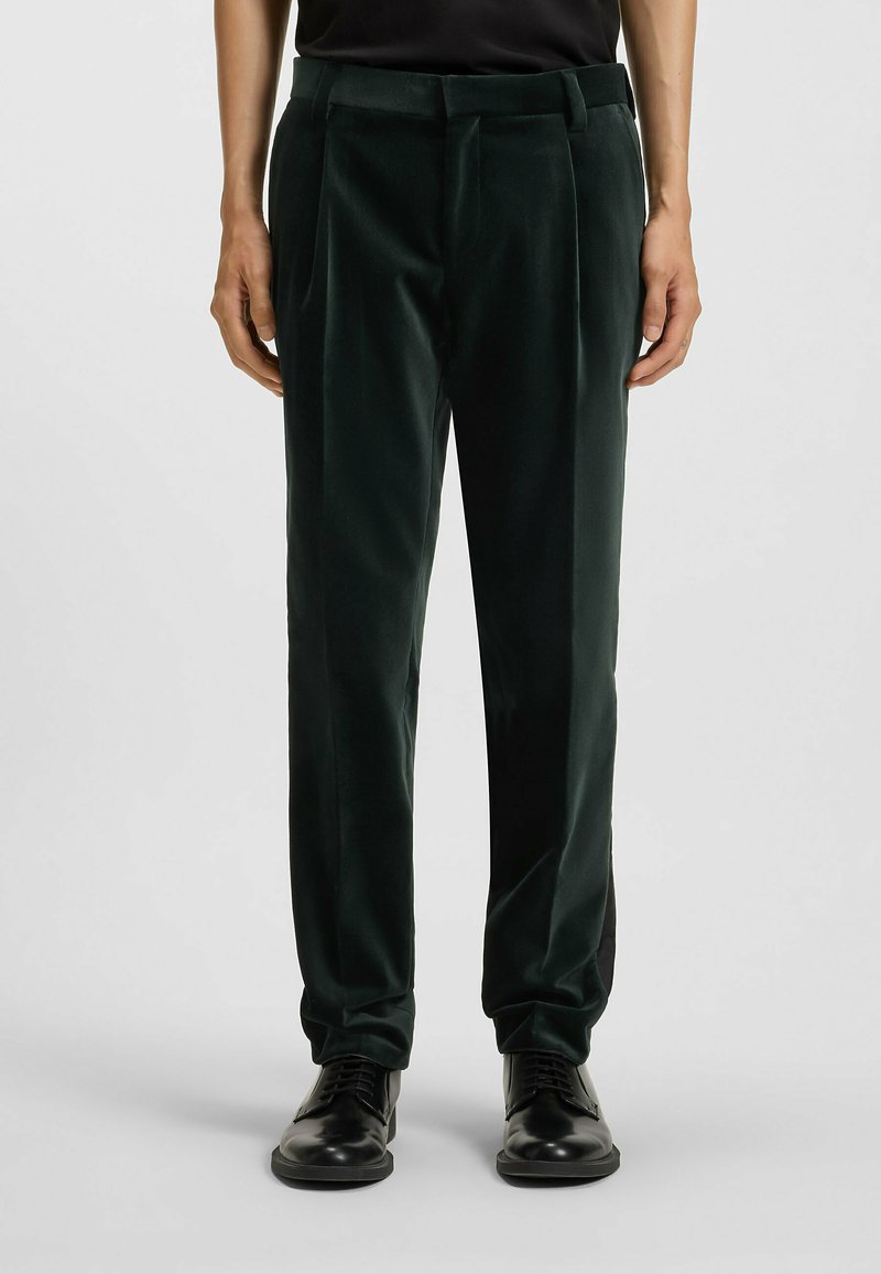 Grüne Samenhosen mit glatter Textur, ausgestattet mit einer taillierten Passform und Falten, kombiniert mit schwarzen formellen Schuhen.