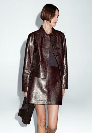 Giubbotto in pelle bordeaux con design corto, bottoni frontali e tasca sul petto. Indossato con una gonna coordinata, creando un look armonioso.