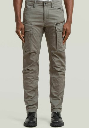 Mann trägt graue Cargohosen mit mehreren Taschen und schwarze Schnürstiefel, steht vor einem neutralen Hintergrund.