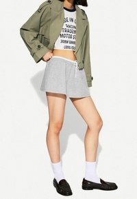 Kobieta w oliwkowej krótkiej kurtce, białym t-shircie z grafiką, szarych szortach, białych skarpetkach i czarnych mokasynach stoi na tle białej ściany.