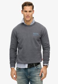 Szary sweatshirt z okrągłym dekoltem i ściągaczami, zdobiony jasnoniebieskim logo "Superdry" na piersi, nałożony na jasnoniebieską koszulę w paski.
