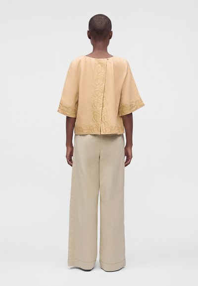 Personne aux cheveux courts, debout, de dos, portant un haut beige ample brodé et un pantalon large beige clair.