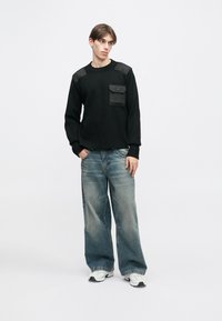 Melns džemperis ar polsterētām plecu daļām un kabatu. Kombinēts ar izbalējušām platajām ziliem džinsiem un baltiem sporta apaviem. Vienkāršs, mūsdienīgs dizains.