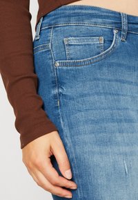 Světle modré džíny z denimu s vybledlým povrchem, které mají malou přední kapsu, měděné kování a detailní záběr rukávu hnědé košile s dlouhým rukávem.