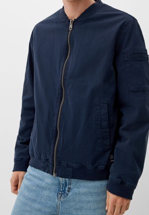 Mężczyzna noszący granatową bomberkę na zamek z prążkowanymi mankietami i dołem, w połączeniu z jasnoniebieskimi jeansami, stojący z rozluźnionymi rękami.