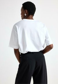 Hvid bomulds t-shirt med korte ærmer, vid pasform, rund hals. Bær med høj talje sorte bukser med baglommer og en glat overflade.