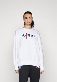 Biely sveter s nápisom "PARÍŽ" v námorníckej modrej a červenej grafike, s okrúhlym výstrihom, voľným strihom a zúženými manžetami, kombinovaný s tmavými nohavicami.