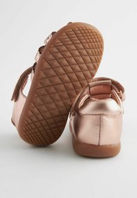 Różowo-złote metaliczne buty dziecięce z teksturowaną gumową podeszwą, wyposażone w rzep i miękkie zapięcie z tyłu ułatwiające zakładanie.
