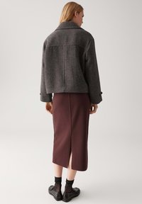 Grijze wollen cropped jas met een brede kraag over een bordeaux midi rok met een split aan de achterkant, gecombineerd met zwarte sokken en schoenen.