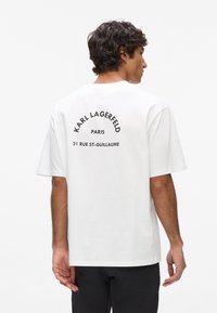 Biela bavlnená tričko s krátkym rukávom, na zadnej strane s čiernym zakriveným potlačeným textom: "Karl Lagerfeld Paris 21 Rue St-Guillaume."
