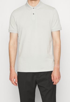 Mann in einem hellgrauen kurzärmeligen Poloshirt mit zwei Knöpfen und schwarzen Hosen, stehend vor einem weißen Hintergrund.