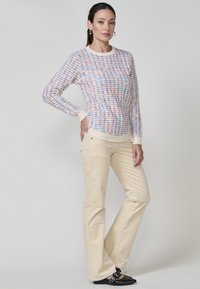 Pull en laine multicolore avec un motif texturé, base crème claire et poignets côtelés, associé à un pantalon beige évasé et des chaussures noires.