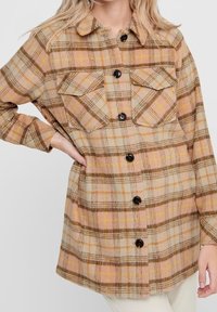 Veste-chemise à carreaux marron et beige ; comporte deux poches poitrine, un col arrondi et des boutons noirs sur un tissu doux et texturé.