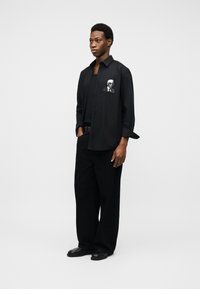 Νεαρός άνδρας ντυμένος εξ ολοκλήρου στα μαύρα, με άνετο πουκάμισο με κουμπιά που φέρει ένα μονοχρωματικό πορτρέτο στο ύψος της τσέπης στο στήθος.