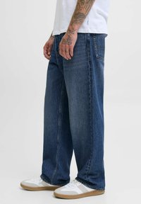 Jean en denim large de couleur bleu foncé avec un design classique à cinq poches, associé à des baskets blanches avec une semelle beige et des accents gris.