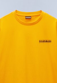Žluté bavlněné tričko s kulatým výstřihem a krátkými rukávy, s potištěným logem v černé barvě na levé části hrudi.