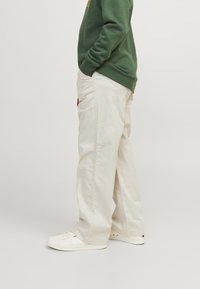 Pantaloni ampi color beige chiaro con tasche laterali, abbinati a una felpa verde. Sneakers bianche dal design minimal. Materiale in cotone morbido visibile.