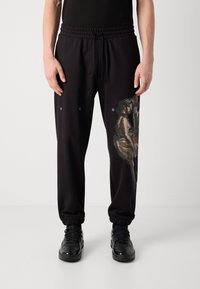 Black sweatpants feature an elastic waistband with drawstring, tapered cuffs, and a large artistic graphic on the left leg. Brand logo present.