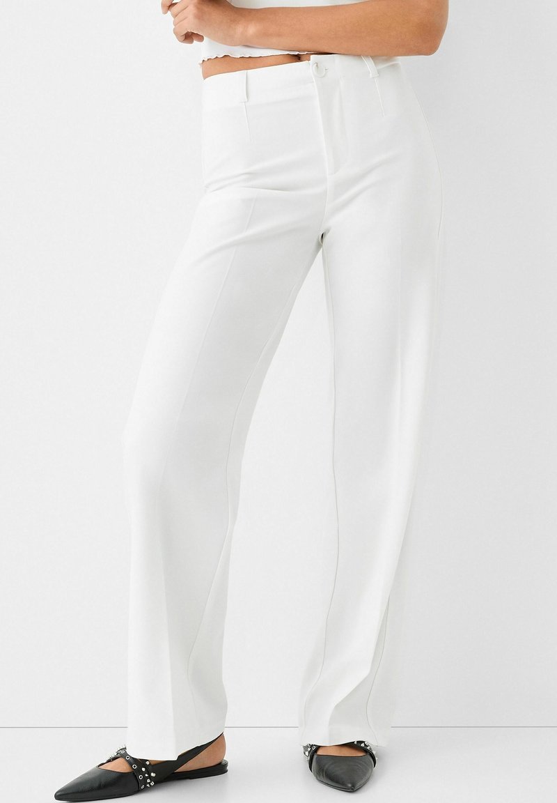 Femme portant un pantalon blanc taille haute à jambes larges et des ballerines slingback noires pointues avec des clous argentés, debout sur un fond blanc.