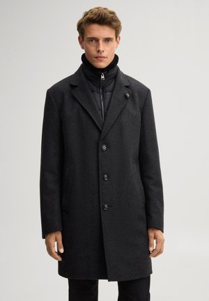 Mann trägt einen schwarzen Dreiknopf-Übermantel über einer gesteppte schwarze Jacke mit Reißverschlusskragen, steht vor einem schlichten hellen Hintergrund.