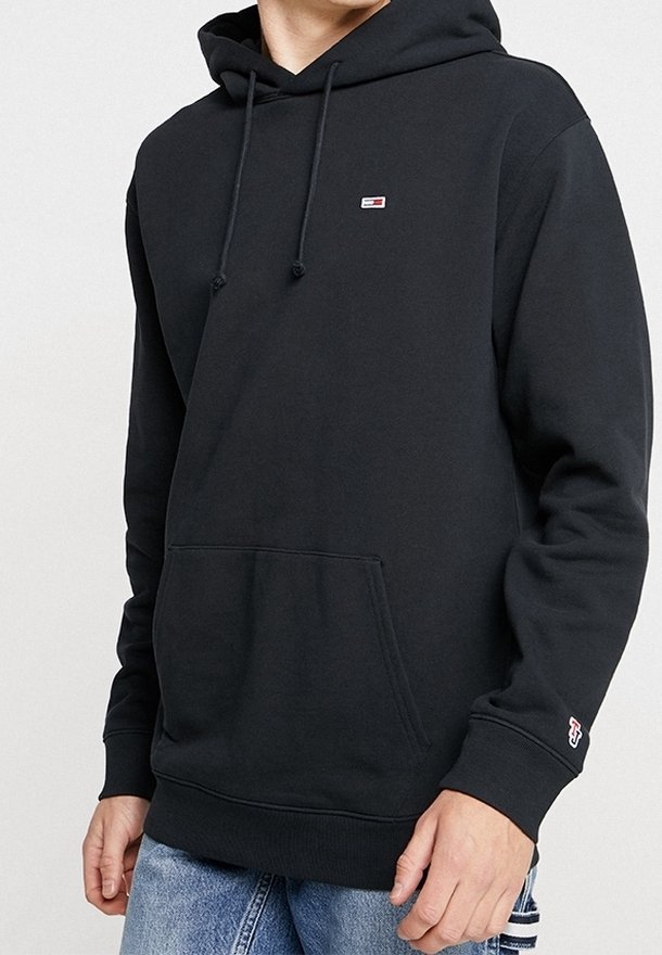 Sudadera negra con cordones, bolsillo tipo canguro, pequeño logotipo de Tommy Hilfiger en el pecho y logotipo "TH" en el puño izquierdo, combinada con jeans azules.