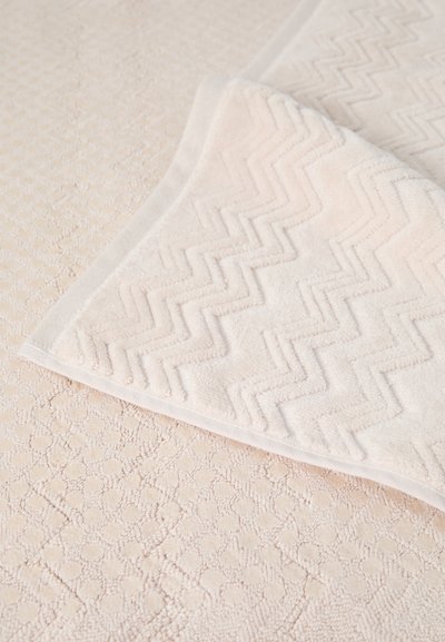 Toalha branca com um padrão em ziguezague no canto superior, texturizada com laços macios. O fundo apresenta um padrão pontilhado em uma cor clara.
