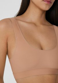 Bezešvá podprsenka v béžové barvě s širokými ramínky, hladkým povrchem a kulatým výstřihem. Disponuje podpůrným podbandem a jemným švem.
