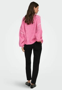 Roze gebreide trui met een losse pasvorm, textuurafwerking en witte stiksels op de mouwen. Gecombineerd met zwarte aansluitende jeans en versierde schoenen.