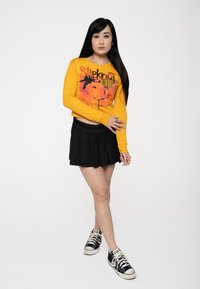 Yellow long-sleeve top with graphic print, paired with a black pleated mini skirt and black sneakers. Adjusted fit and casual design.