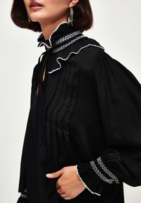 Schwarze Bluse mit Rüschkragen und strukturiertem Stoff; verfügt über vertikale Falten und kontrastierende weiße Nähte an den Manschetten und am Kragen.