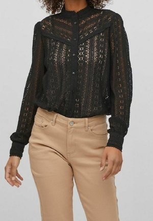 Femme portant un chemisier noir en dentelle transparente à boutons, manches longues, et un pantalon beige taille haute, sur un fond uni.