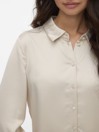 Lichtbeige satijnen blouse met een puntige kraag, voor gesloten knopen en lange mouwen. Tedere textuur, minimalistisch ontwerp, geen patronen.