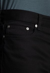 Jeans de mezclilla negra con una textura distintiva, que cuentan con bolsillos frontales, detalles de botones plateados y una vista en primer plano de la cinturilla.
