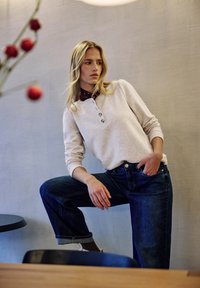 Jasnoszara bluza z guzikami, zestawiona z ciemnoniebieskimi dżinsami. Modelka pozuje na tle teksturowanej ściany z rozmytym tłem.