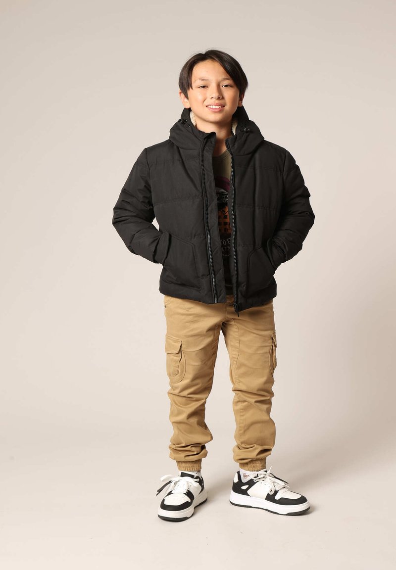 Doudoune noire avec capuche, fermeture éclair à l'avant et deux poches ; associée à un pantalon cargo beige et des sneakers noir et blanc. Fond neutre.