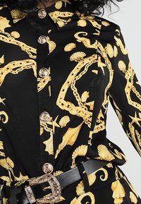 Svart skjorta med ett mönster av gula snäckor, kedjor och havsdjur; har guldfärgade knappar och ett svart bälte med en utsmyckad spänne.