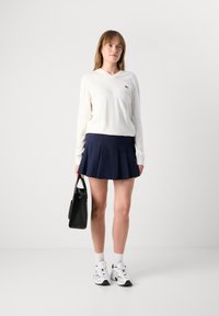 Hvit V-hals genser, marinblå, plisserte skjørt, svart håndveske, hvite sokker og hvite/svarte joggesko. Enkel design, myk tekstur og ledig passform.
