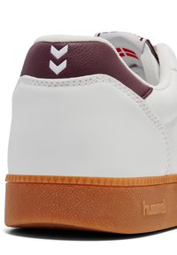 Witte sneaker met maroon leren hiel, gestructureerde gumrubberen zool, witte steken en chevronlogo-detail op de achterkant.